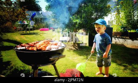 Young boy looking at smoking barbecue in a summer garden - John Gollop Stock Photo