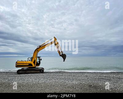 Excavator working on earthmoving at open pit mining on sea and sky clouds background. Backhoe digs sand and gravel in quarry. Heavy construction equip Stock Photo