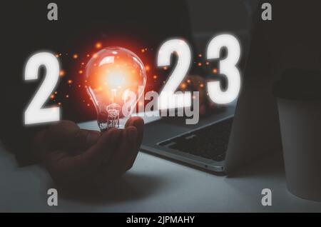 Business hand holding bulb light text 2023 and computer laptop on desk.Idea innovation growth plan concept. Stock Photo