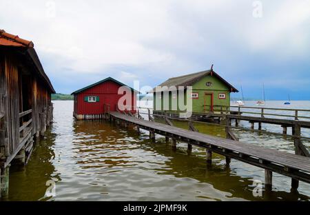 a long wooden pier leading to the colorful boat houses on lake Ammersee with sailing boats and the Alps in the background in the Bavarian Schondorf Stock Photo