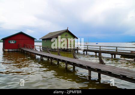 a long wooden pier leading to the colorful boat houses on lake Ammersee with sailing boats and the Alps in the background in the Bavarian Schondorf Stock Photo