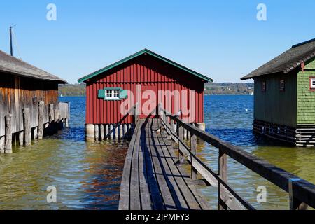 a long wooden pier leading to the colorful boat houses on lake Ammersee with sailing boats and the Alps in the background in the Bavarian Schondorf Stock Photo