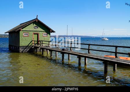 a long wooden pier leading to the colorful boat houses on lake Ammersee with sailing boats and the Alps in the background in the Bavarian Schondorf Stock Photo