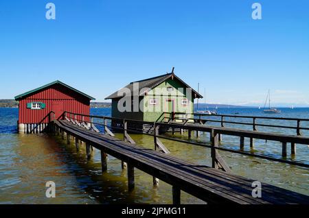 a long wooden pier leading to the colorful boat houses on lake Ammersee with sailing boats and the Alps in the background in the Bavarian Schondorf Stock Photo