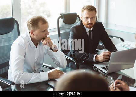 A group of company employees sits at a table in a meeting room. A team of young businessmen working and communicating together in an office. Corporate Stock Photo