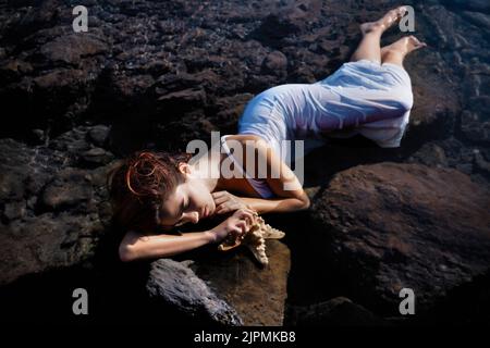 From above of feminine young woman with long dark hair in wet white dress lying on rough boulders with closed eyes near starfish at seaside Stock Photo