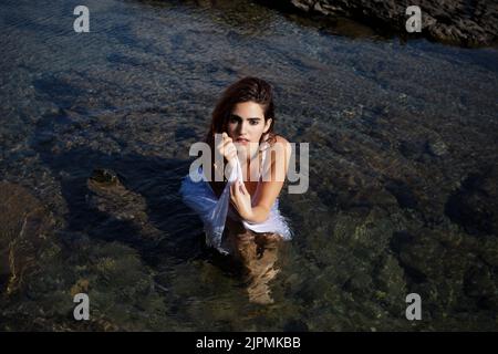 From above of confident young female model with long brown hair in white dress sitting in shallow water of sea with rocky bottom and looking at camera Stock Photo