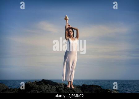 Full body of gorgeous young barefooted female in summer white maxi dress standing on rocky formation with raised arms under cloudy blue sky near sea Stock Photo