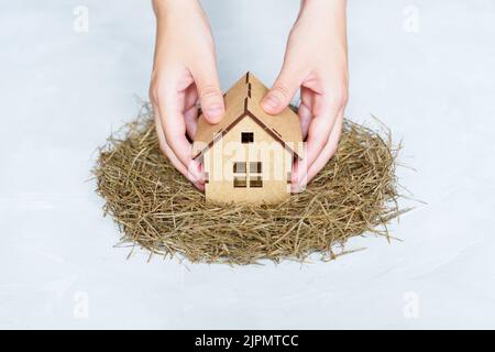 Placing a miniature wooden house in a hay nest holding it with both hands isolated on gray background. Creative family home concept. Stock Photo