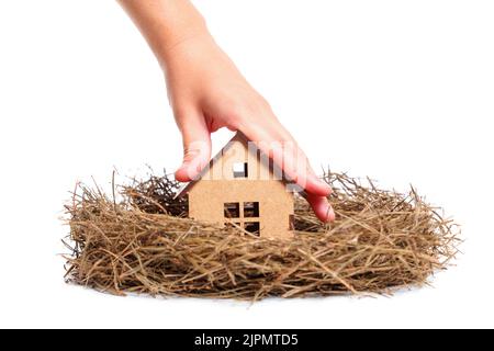 Hand placing a miniature wooden house in a hay nest isolated on white background. Family house concept. Stock Photo