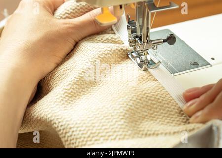 Seamstress hands holding white textile fabric. Female hands stitching white fabric on modern sewing machine at workplace. Close up view of sewing proc Stock Photo