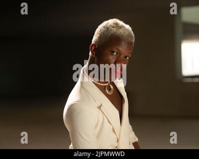Stylish African American male model in elegant white jacket with short blond hair and red lips looking at camera with serious face on blurred backgrou Stock Photo