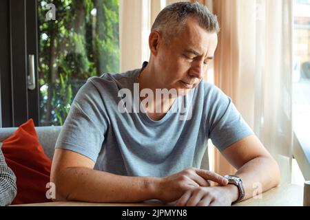 Portrait of thoughtful elderly man sitting at table on sofa near window, looking at watch, waiting for meeting. Date. Stock Photo