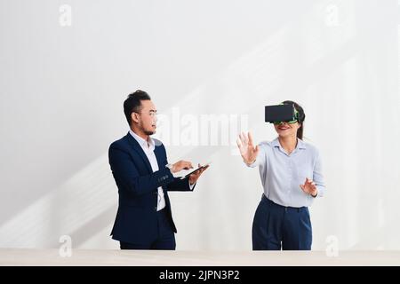 Cheerful young developer in formalwear using VR headset while testing new game with handsome Asian colleague, white background Stock Photo