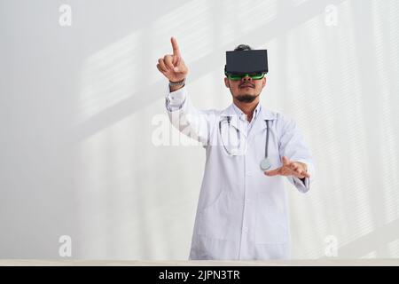 Concentrated bearded doctor wearing white coat pointing at something while using VR headset at modern office, white background Stock Photo
