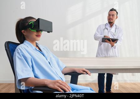 Smiling young nurse sitting on cozy chair and using VR headset while confident bearded therapist carrying out experiment, interior of modern office on background Stock Photo
