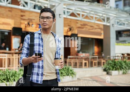 Portrait shot of handsome Asian man looking away pensively while listening to music in headphones and enjoying fragrant coffee, modern outdoor cafe on background Stock Photo