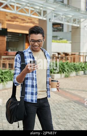Full length portrait of smiling Asian man in headphones browsing Internet on smartphone while enjoying fresh air and delicious cappuccino outdoors Stock Photo