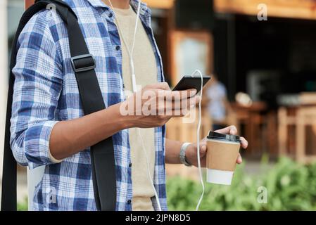 Close-up shot of unrecognizable man wearing T-shirt and checked shirt surfing Internet on smartphone and drinking fragrant coffee while standing outdoors, blurred background Stock Photo