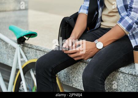 Close-up shot of young cyclist using smartphone while sitting on marble bench and enjoying fragrant coffee Stock Photo