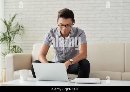 Portrait shot of smiling Asian student sitting on cozy sofa of modern living room and preparing for exams with help of laptop Stock Photo