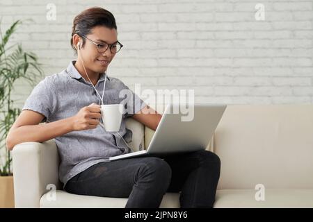 Handsome young freelance worker sitting on cozy sofa with cup of fragrant coffee and working on promising project, interior of modern living room on background Stock Photo
