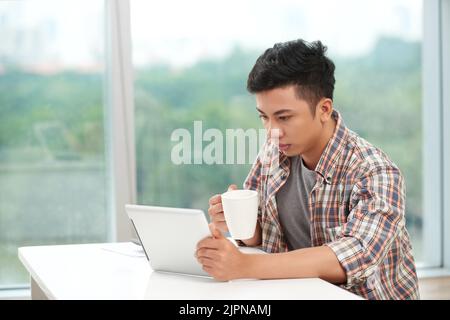 Young Asian man driking morning coffee and reading news on digital tablet Stock Photo