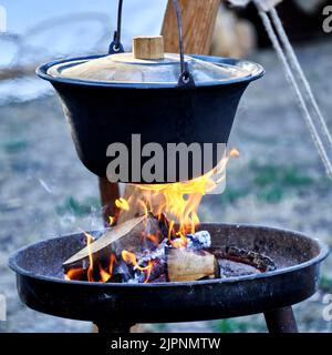 Cast iron pot with primitive lid hangs over open fireplace with burning logs Stock Photo