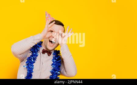Funny guy 20-25s in a party hat and New Year's tinsel laughs and makes a gesture with his hands in the form of glasses or binoculars, in the form of a Stock Photo
