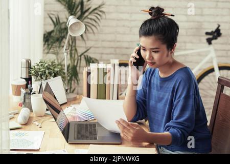 Young Vietnamese graphic designer reading document and calling to client Stock Photo