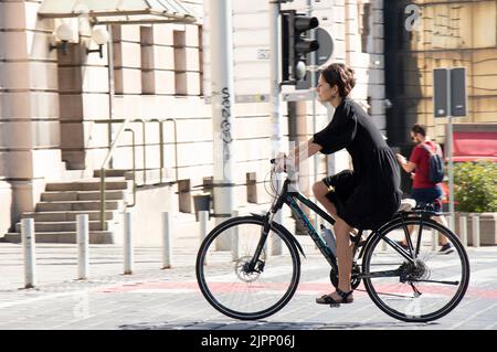 Belgrade-Serbia - June 15, 2022: Elegant young brunette woman wearing black dress riding a bike on city street Stock Photo