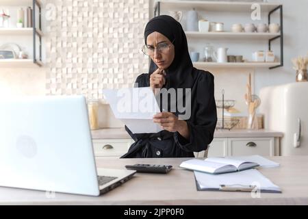 Arab woman at home doing paperwork, concentrated Muslim woman in hijab holding bills in hands, calculating household budget paying loan, using laptop in kitchen at home Stock Photo