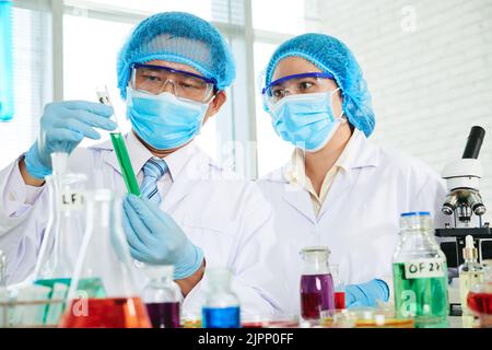 Concentrated Asian scientists wearing white coats and medical masks examining liquid in test tube while gathered together at modern laboratory Stock Photo