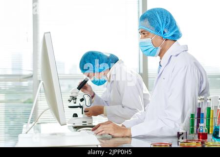 Team of talented microbiologists wearing white coats and medical masks wrapped up in work at modern lab: Asian woman studying sample with help of microscope while her male colleague taking notes Stock Photo