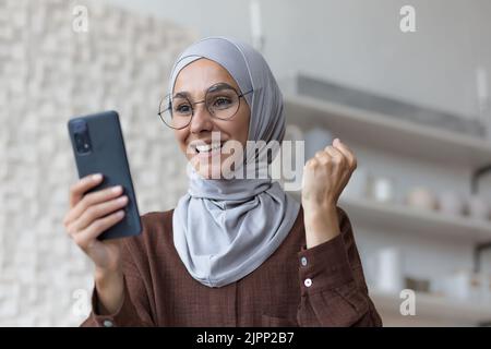 Young beautiful arab woman using phone smiling and celebrating success, muslim woman in hijab in living room holding smartphone Stock Photo