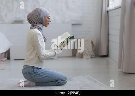 A young beautiful Muslim woman in a hijab is praying at home, sitting by the window Stock Photo