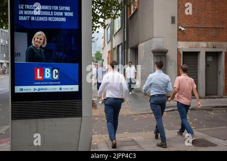 Lunchtime workers walk past a news headline for Conservative leadership contender, Liz Truss who is seen on a digital media screen, on 17th August 2022, in London, England. A leaked conversation shows Truss as saying that British workers are lazy and in leaked comments, needing 'more graft.' Stock Photo