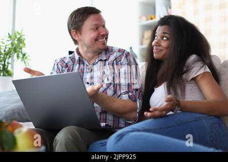 Interethnic family couple laughing and chatting while sitting on sofa at home with laptop. Stock Photo