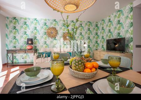 Dining room with wooden table with large breakfast area with fruits, juices and assorted pastries, living room with furniture and decoration on the wa Stock Photo