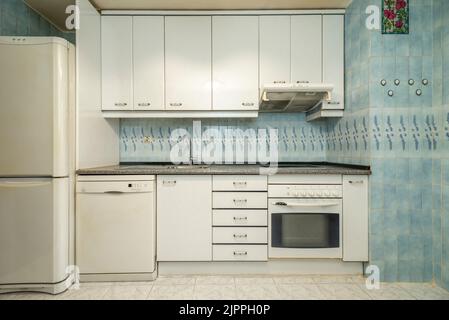 Front view of a kitchen with white wood cabinets, built-in appliances, pink granite countertops, and blue tile on the walls. Stock Photo