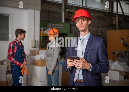Portrait of an elegant engineer with protective helmet and tablet inside the construction site with workers behind. Stock Photo