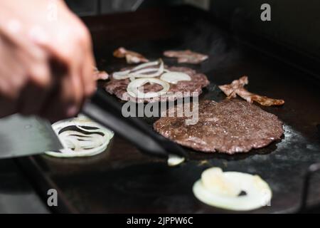 close-up detail shot of a man grilling two ground hamburger meats with onions and bacon around them, cooking in a Colombian street food grill or fast Stock Photo