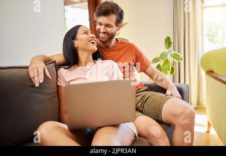 Couple talking and browsing on a laptop while relaxing on a sofa at home. Girlfriend and boyfriend scrolling on social media or the internet with Stock Photo