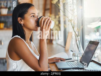 Young freelance worker drinking coffee and working on a laptop in a cafe. Woman entrepreneur reading work or business email online with technology Stock Photo