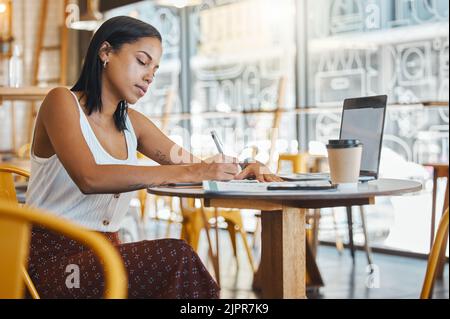 Student writing, taking notes or doing a project assignment in cafe or restaurant using laptop and notebook. Young woman taking an online education Stock Photo
