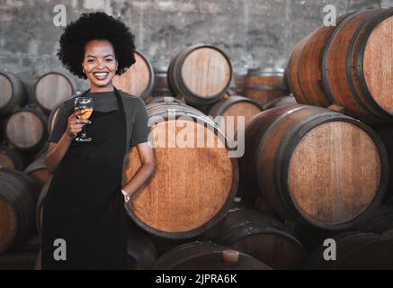 Portrait of a young woman winemaker standing with a glass with wooden barrel of red wine in a winery cellar or distillery. Entrepreneur or business Stock Photo