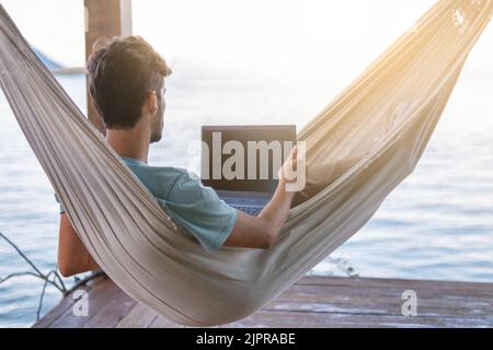 young digital nomad lying on hammock with sea views working remotely on internet at sunset - Traveling with a computer - Dream online job concept - Se Stock Photo