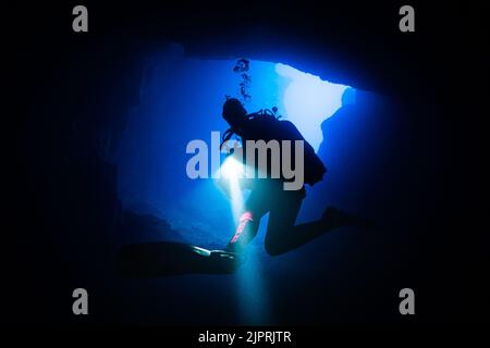 Diver turning around in front of a cave exit with shining flashlight in hand, while light shines in from outside in Blue Hole on Gozo, Malta. Stock Photo