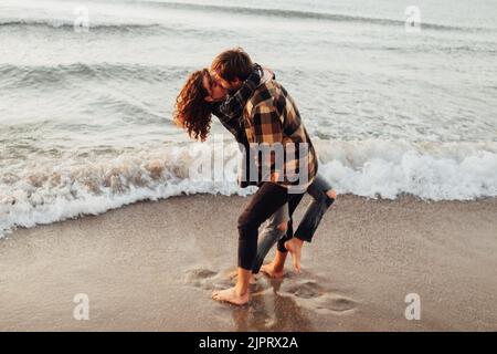 Young woman and man hugging and kissing together while standing on the seashore at dawn Stock Photo