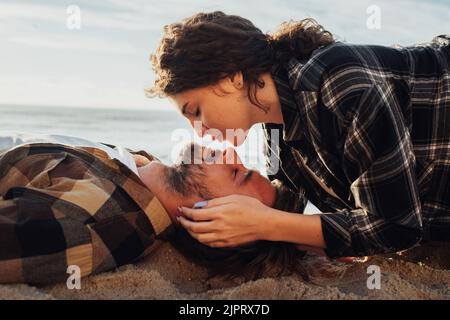 Young adult couple laying on the beach of sea, caucasian curly woman kissing her boyfriend Stock Photo